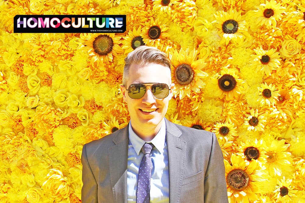 A professional gay man, wearing a suit and sunglasses, in front of a wall of sunflowers.