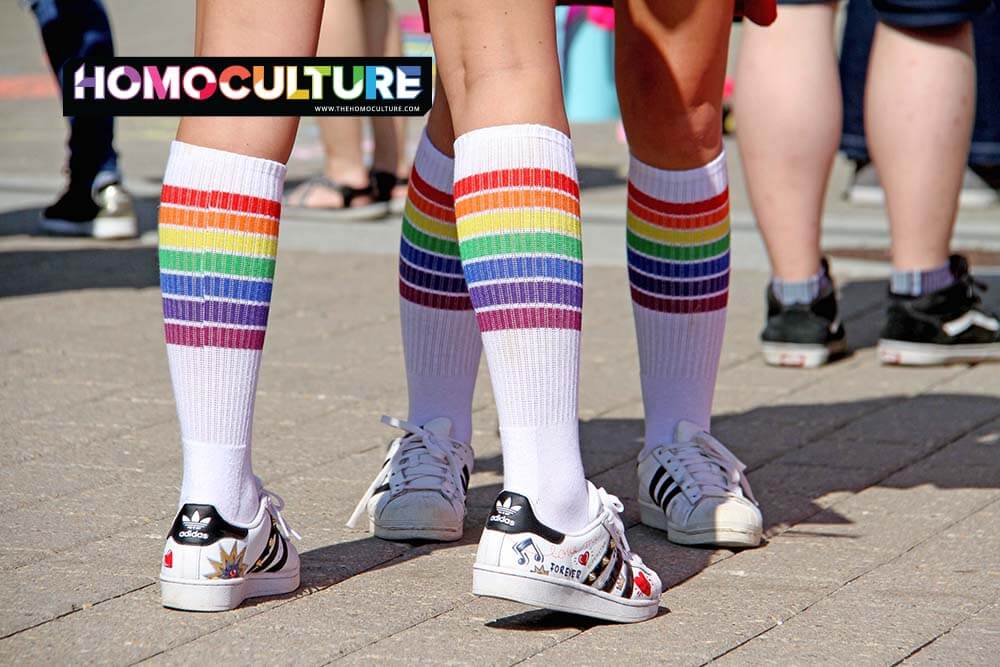 Close-up of two people wearing rainbow-striped knee-high socks and white Adidas sneakers at a Pride event, symbolizing LGBTQ pride and community celebration.