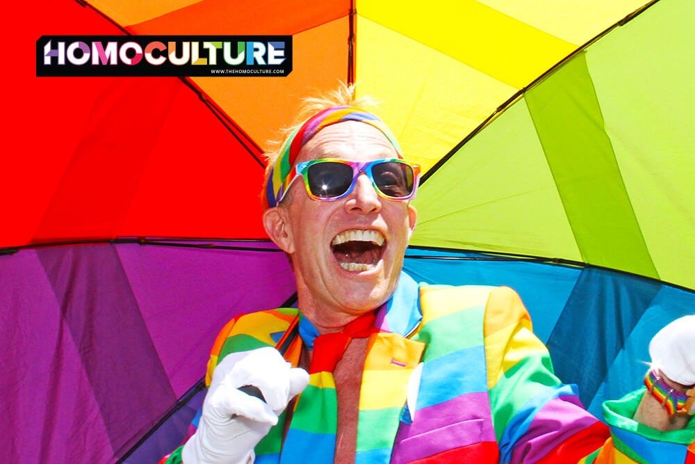 A gay man at a Pride parade dressed in a rainbow outfit, holding a rainbow umbrella.