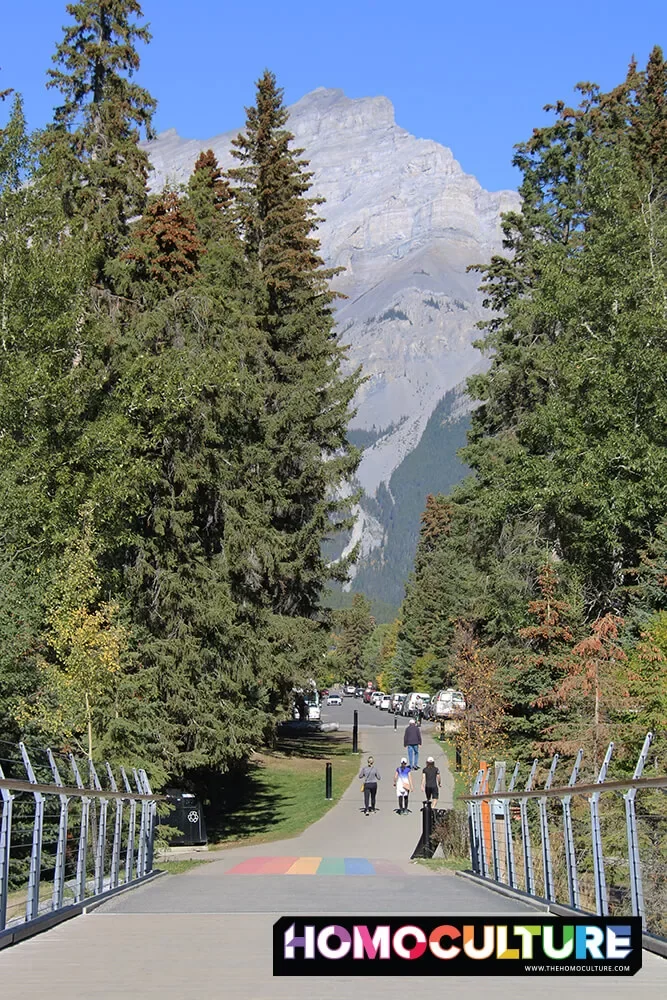 A view of the Pride crosswalk at the pedestrian bridge in Banff, Alberta, with the Rocky Mountains in the background. 
