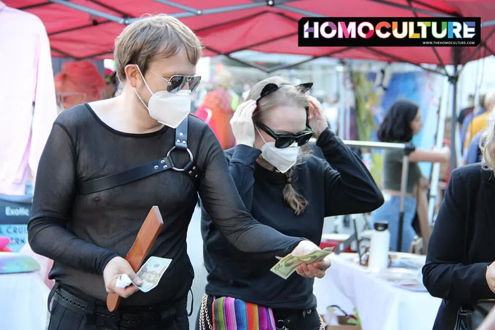 A couple wearing fetish gear and N95 masks buying a wood paddle at Folsom Street Fair 2022.