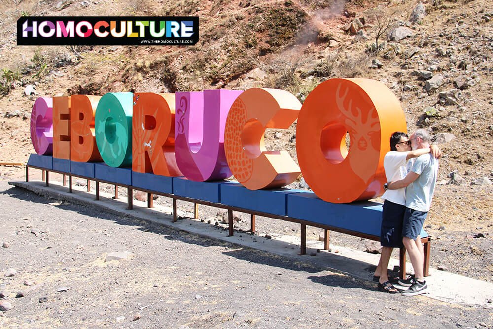 A gay couple kissing in front of the Ceboruco sign.