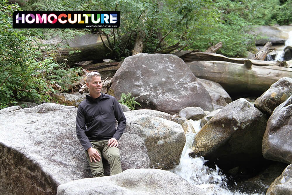 Queer travel safety: a man leaning against a rock near a river in the mountains.