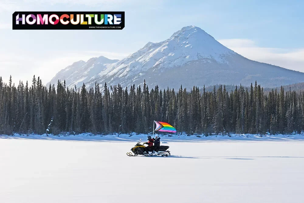 Friends riding a snowmobile on Tagish Lake near Whitehorse on a sunny day, carrying a progressive Pride flag. 