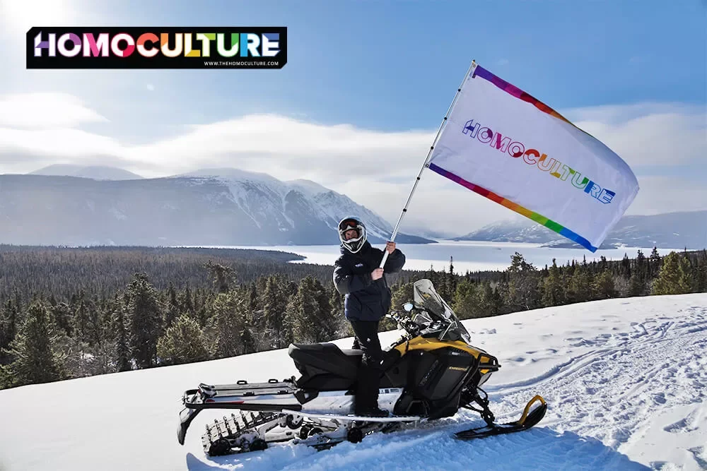 HomoCulture founder, Brian Webb, waves a HomoCulture flag while standing on a snowmobile with the Yukon wilderness in the background.