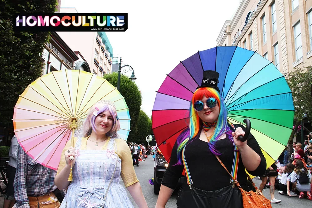 Two girls holding up coloruful umbrellas at a Pride parade.