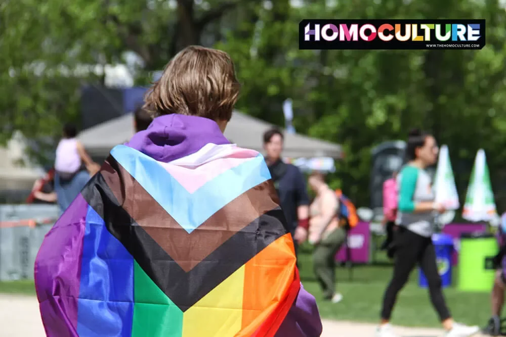A young queer adult wearing a progressive pride flag through a park on a summer day.