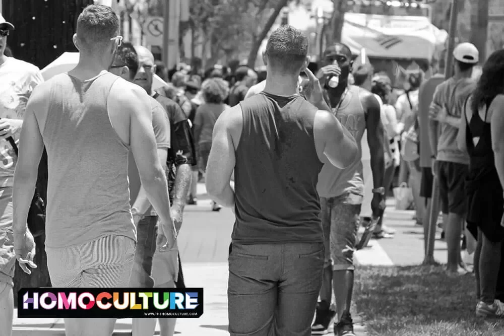 A greyscale image of two guys in tanktops and shorts walking down a busy sidewalk.