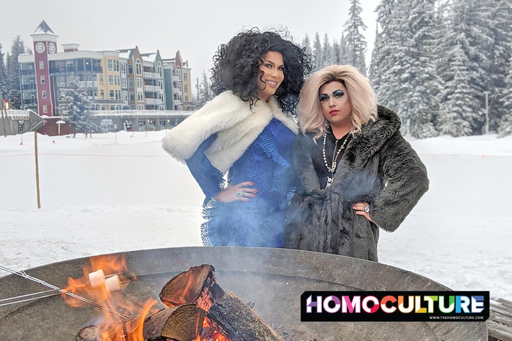 Two drag queens outside in the winter, by a bon fire.