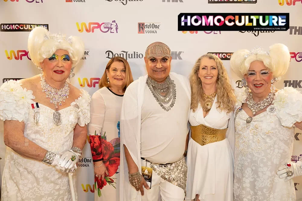 A group of friends in matching outfits stop for a photo on the white carpet at the White Party Palm Springs 2024 Saturday night White Party Sahara Desert Nights party.
