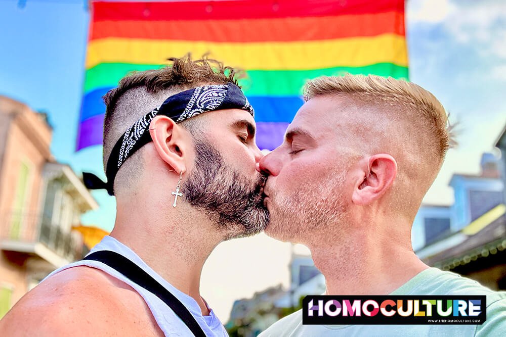 Two gay men kissing in front of a Pride flag.