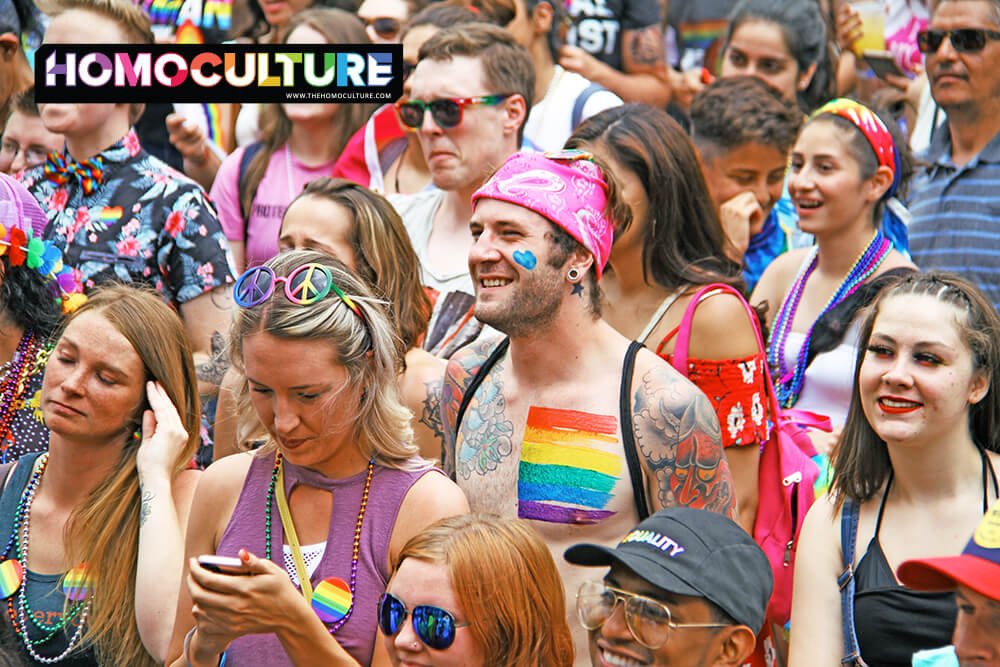 A crowd of people wearing colorful outfits watching a Pride parade.