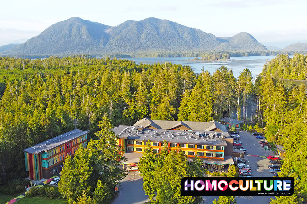 Aerial view of Hotel Zed Tofino, a colorful retro-themed hotel nestled in the lush forest of Vancouver Island, British Columbia. Surrounded by dense trees and coastal waters, the hotel stands out with its vibrant exterior and scenic mountain backdrop.