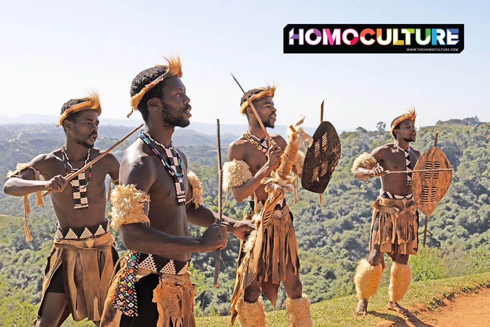 Four Zulu men in traditional warrior attire performing with shields and spears against a scenic hillside backdrop at Phezulu Safari Park.