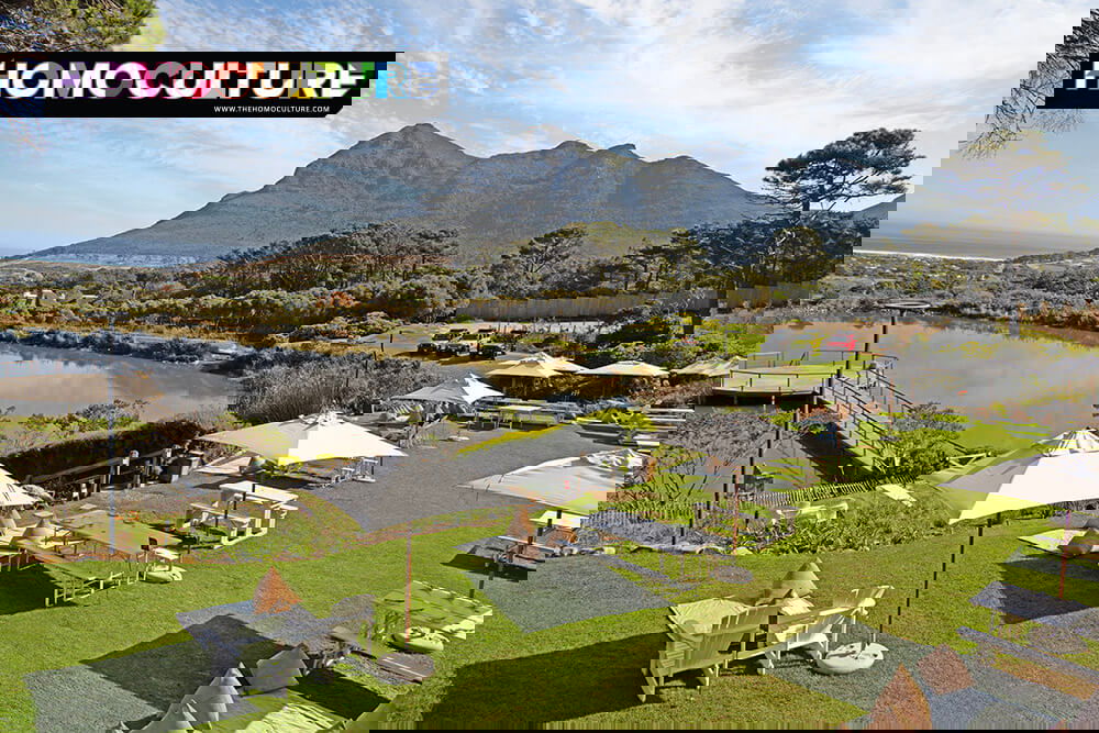 Scenic view of the lawn picnic area at Cape Point Vineyards in Noordhoek, South Africa, with white umbrellas, mountain views, and a reflecting pond near the ocean.