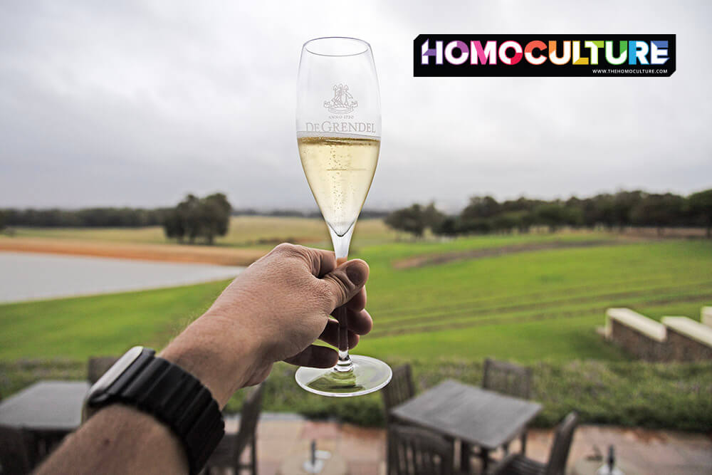 Close-up of a sparkling wine glass held up on the indoor patio at DeGrendel Wine Estate near Cape Town, overlooking the vineyard and farmland on a cloudy day.