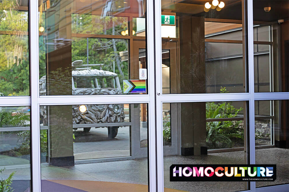 A wooden art car resembling a vintage VW van is reflected in the glass doors of Hotel Zed Tofino. A Progress Pride flag with an intersex symbol is displayed on the entrance, signaling the property's LGBTQ+ inclusivity. Lush greenery and hotel architecture are visible through the reflection.