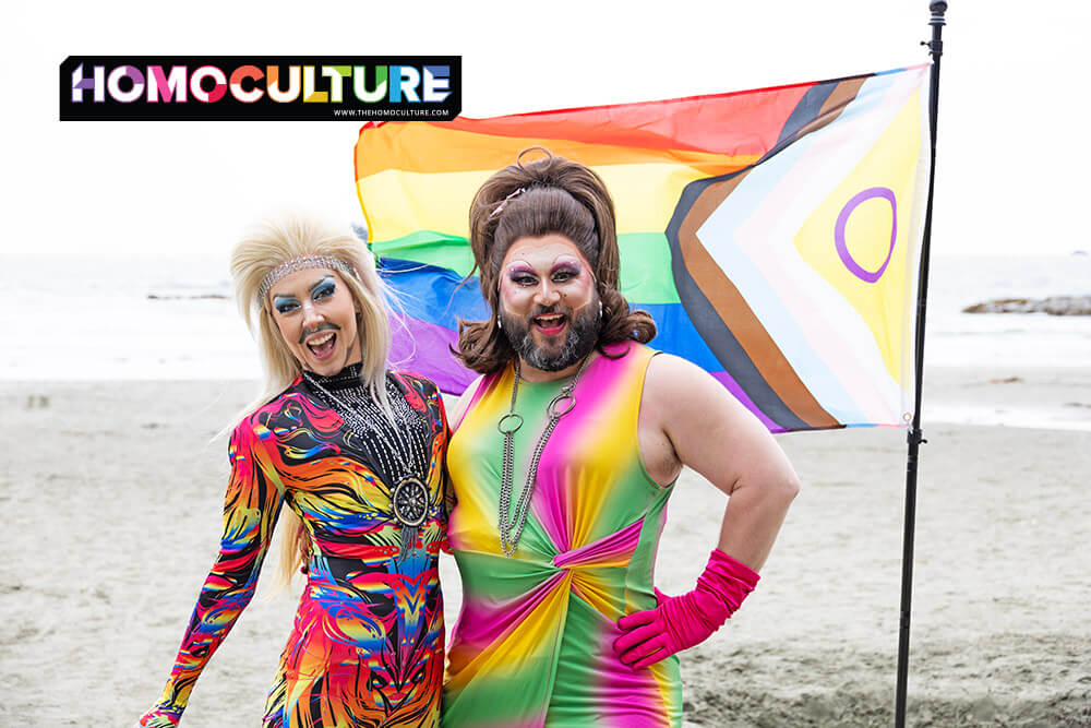 Two drag performers pose joyfully on a sandy beach during Tofino Pride, wearing colorful, eye-catching outfits. Behind them, a Progress Pride flag with the intersex circle waves in the ocean breeze. Both queens smile brightly, radiating Pride spirit.