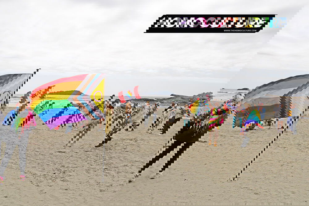A lively crowd gathers on the beach during Tofino Pride, waving Progress Pride and Canadian rainbow flags. Participants in colorful outfits dance and celebrate by the ocean, with cloudy skies and coastal rock formations in the background.