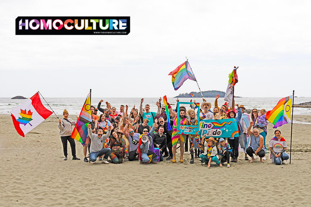 A large, joyful group of people pose together on a sandy beach during Tofino Pride, holding rainbow flags and a colorful “Tofino Pride” banner. Various LGBTQ+ flags, including the Progress Pride flag, intersex flag, and Canadian rainbow maple leaf flag, are proudly displayed. The ocean and small islands appear in the background.