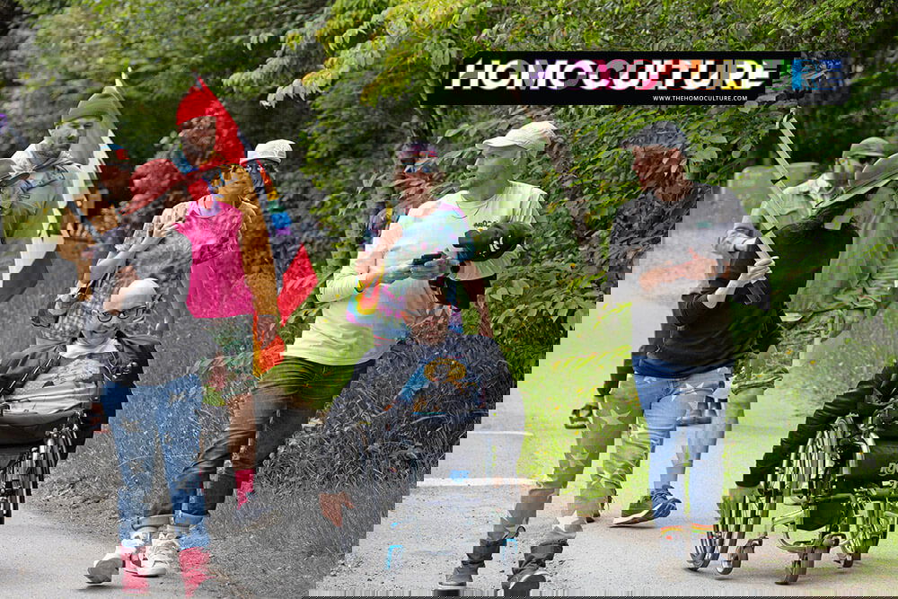 A diverse group of people walk together down a rural road during Tofino Pride, including a person in a wheelchair and others holding flags and wearing rainbow-themed clothing. The group is surrounded by lush greenery, creating an inclusive and joyful atmosphere.