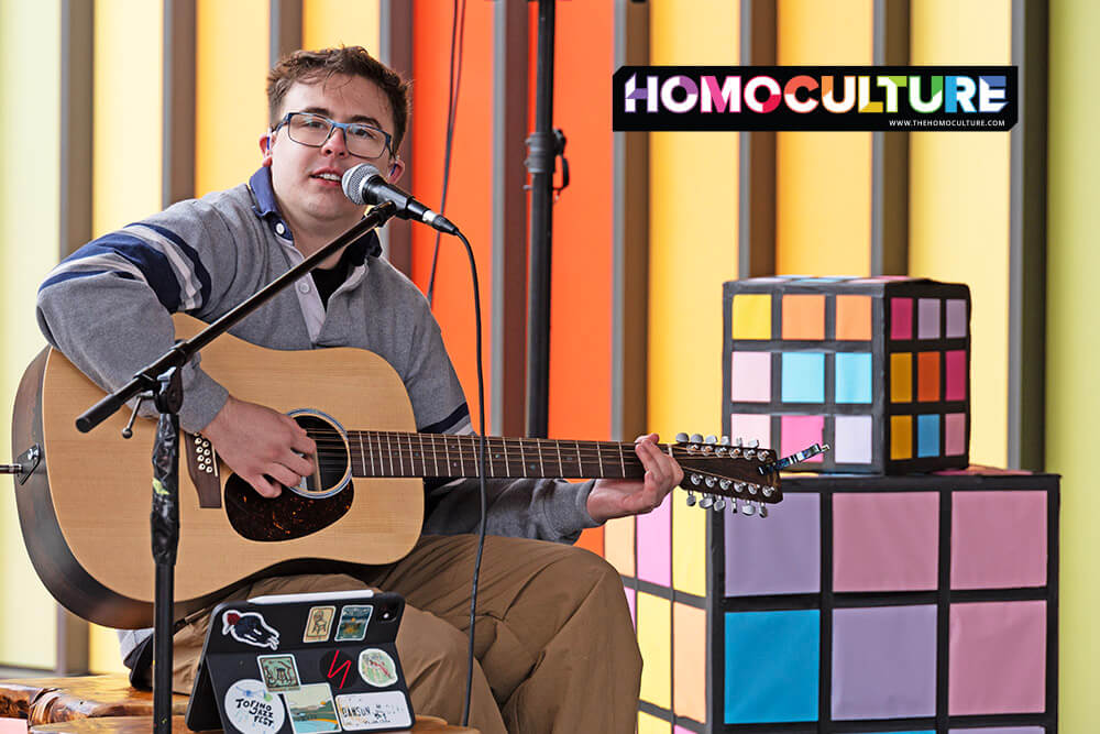 Musician Hayden Roth performs live on acoustic guitar in front of a seated outdoor audience during a Pride event. Colorful hats, rainbow attire, and festive decorations surround the crowd, with a decorated vintage station wagon and rainbow-themed props in the background.