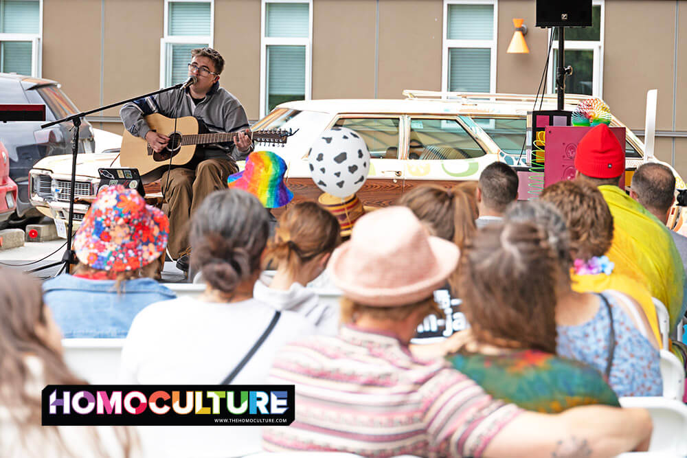 Hayden Roth performs live while playing an acoustic guitar and singing into a microphone. They are seated in front of a rainbow-colored backdrop with a large, cube-shaped art piece painted in bright Pride flag colors. A laptop covered in stickers rests nearby.