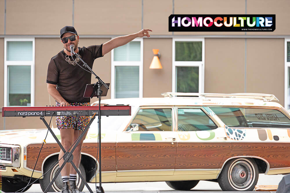 Musician Matthew Presidente performs live at the Tofino Pride festival, singing into a microphone while playing a Nord keyboard. Dressed in a sheer black shirt, patterned shorts, and sunglasses, he gestures to the crowd with one arm. A colorful vintage station wagon is parked behind him.