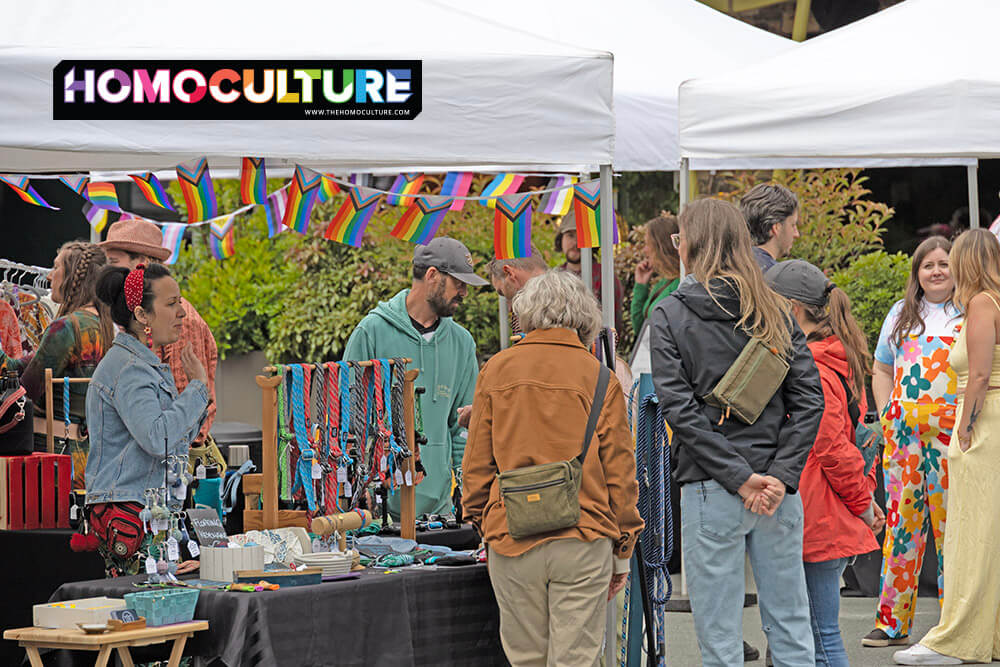 Shoppers explore the Vendor Village at the Tofino Pride Festival, browsing handmade goods, accessories, and crafts under white market tents. Rainbow bunting and Pride flags decorate the booths, creating a festive and inclusive atmosphere. Vendors and attendees chat and browse in colorful, casual attire.