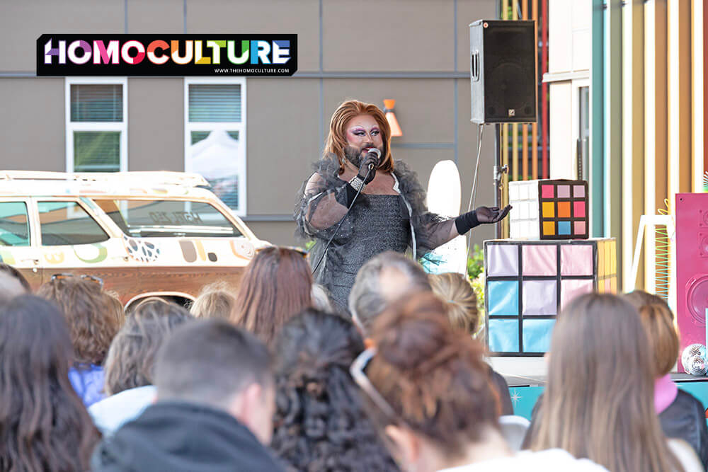 Drag performer Woofie captivates the audience with a microphone in hand during a Tofino Pride show, dressed in a sparkly black outfit with dramatic makeup and accessories. A colorful Rubik’s cube-style prop and a decorated vintage station wagon enhance the lively outdoor stage.