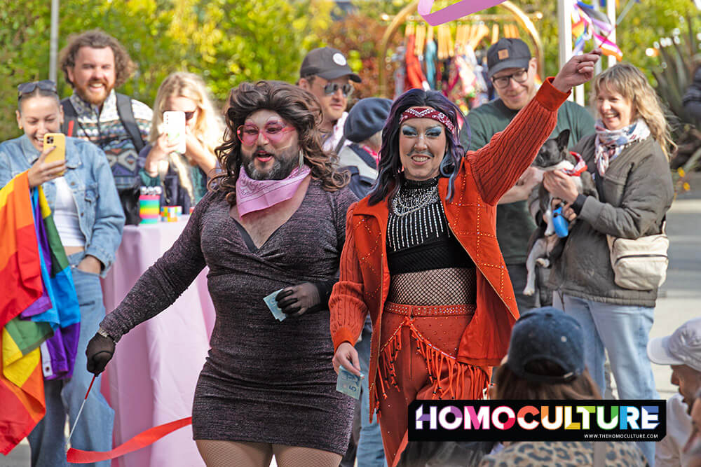 Drag performers Woofie and Eddi-licious dazzle the crowd during a lively performance at the Tofino Pride festival. Surrounded by cheering spectators, rainbow flags, and flashing cameras, the duo struts confidently—Woofie in a glittery dress and pink sunglasses, and Eddi-licious in a bold red fringed outfit.