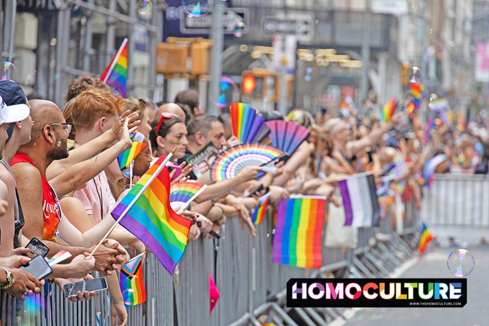 Crowd of LGBTQ+ supporters lined up behind barricades waving rainbow Pride flags and handheld fans at the NYC Pride March, celebrating queer visibility, love, and community in the streets of Manhattan.