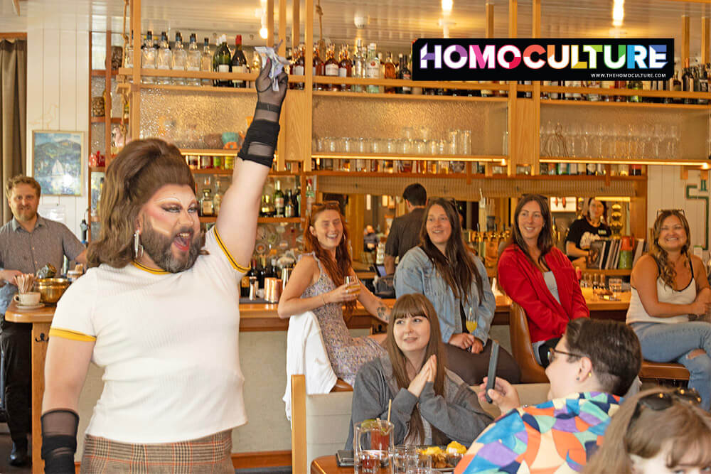 Drag performer Woofie entertains a cheerful crowd at the Tofino Pride drag brunch, holding cash tips in the air with a big smile. Surrounded by delighted guests in a cozy restaurant setting, Woofie wears a retro-style outfit with dramatic makeup and jewelry.