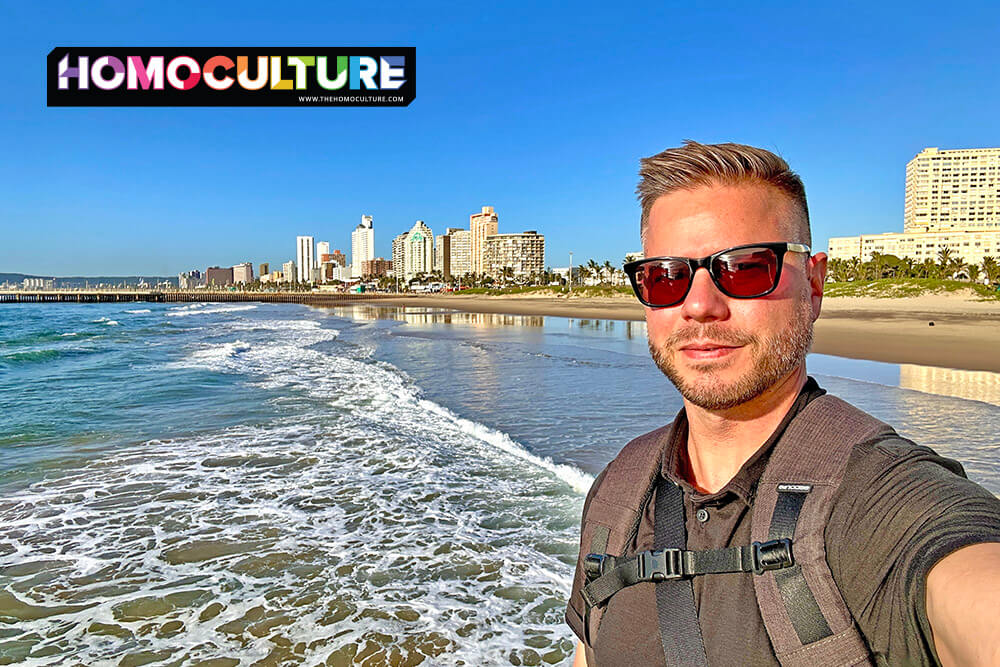 Man wearing sunglasses and backpack taking a selfie on the beach with Durban’s skyline and surf in the background.