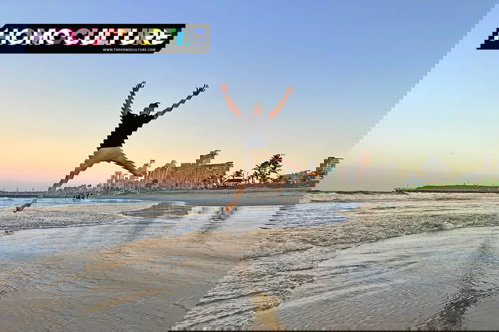 A gay traveler leaps into the air on Durban’s Golden Mile beach at sunset, with the city skyline glowing in the background and soft ocean waves at his feet.