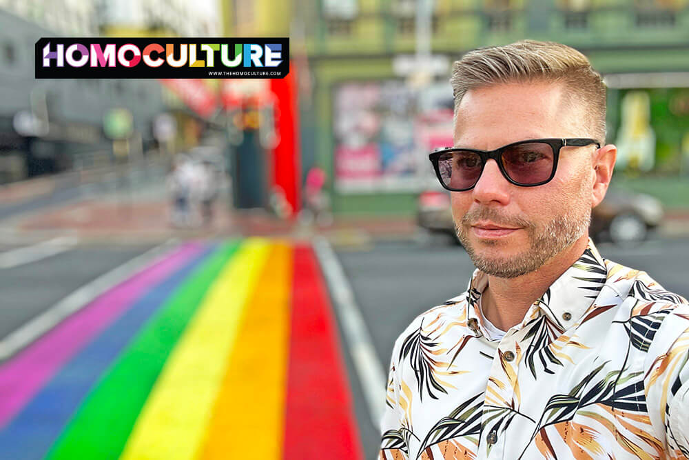Man wearing sunglasses and a floral shirt taking a selfie at a rainbow-painted crosswalk in Cape Town, South Africa.