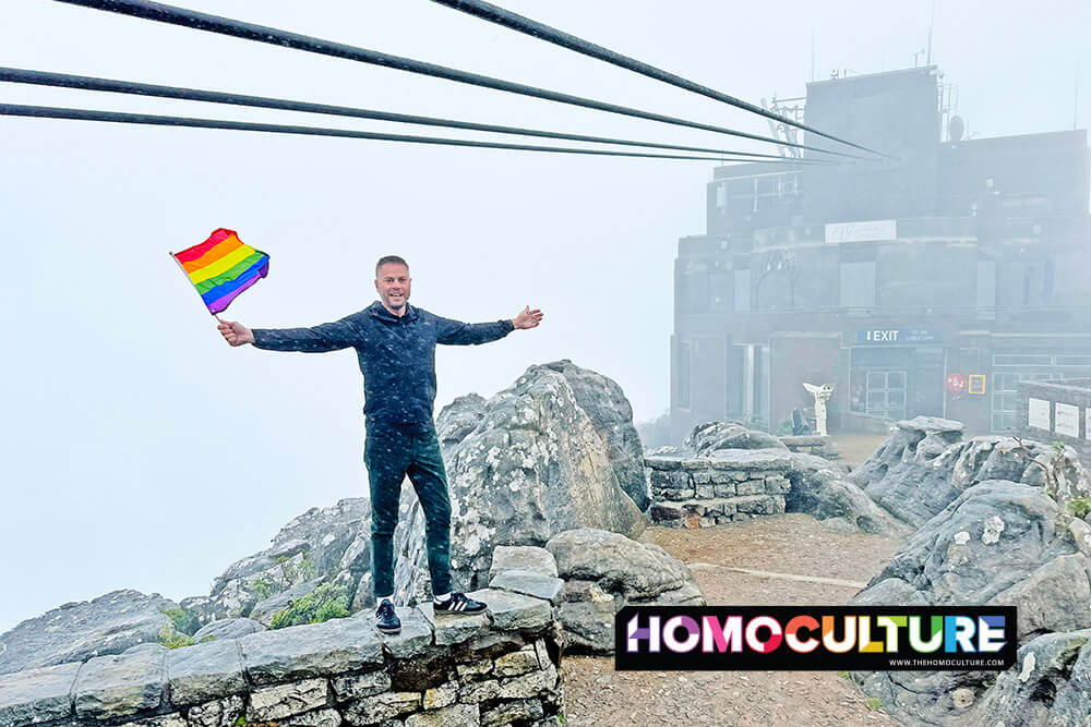 Man standing on a rocky ledge waving a rainbow Pride flag in the mist at the summit of Table Mountain in Cape Town.